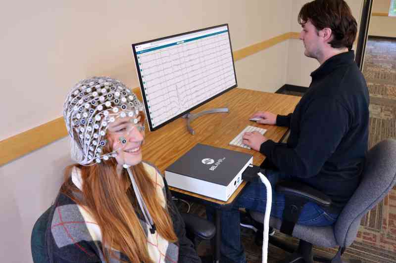 A young woman wearing a BEL Geodesic Head Web sits next to a table where a technician is using a computer connected to a BEL AMP One.